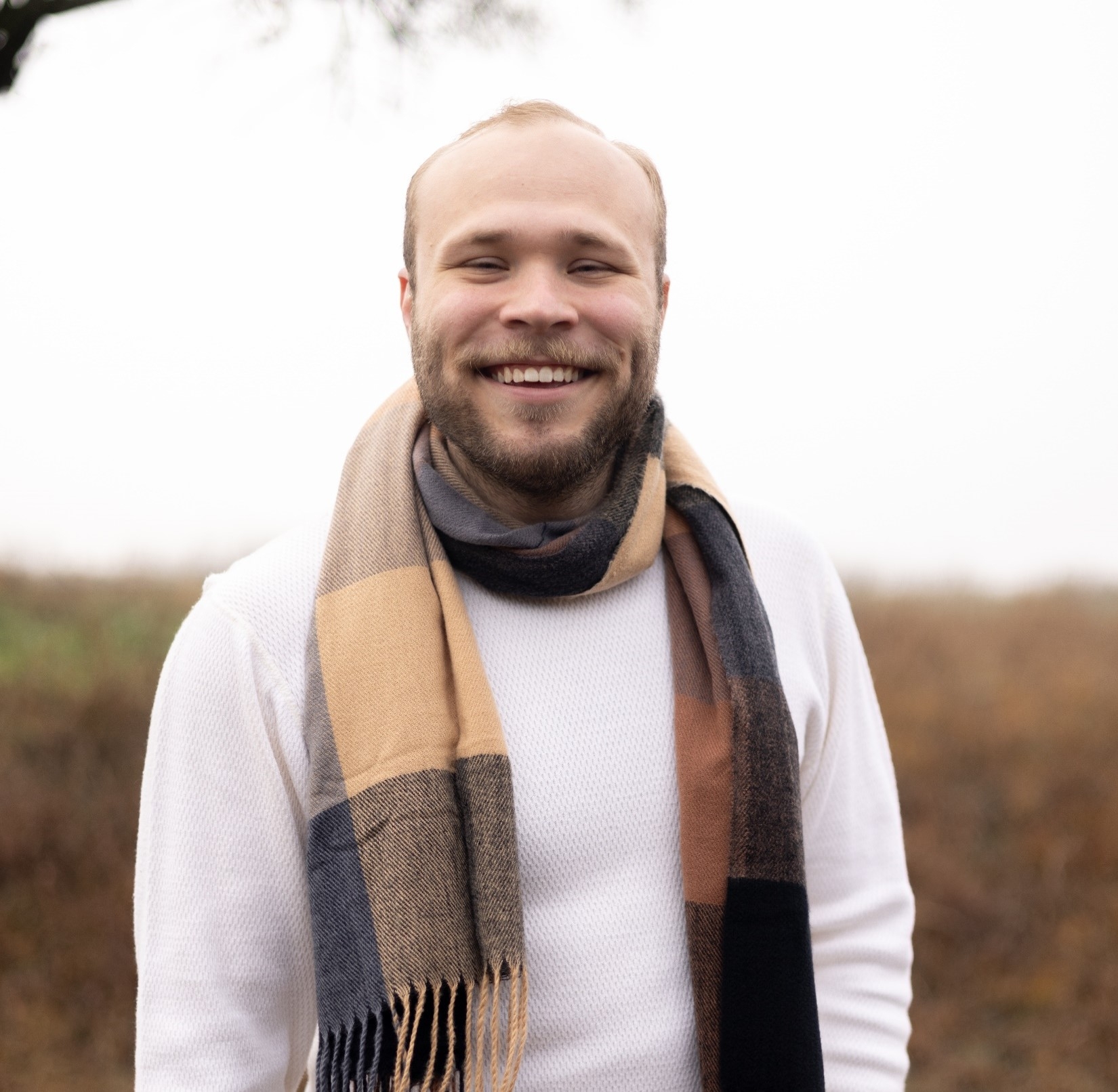 Headshot of Alexander J. Edwards on a foggy day in the suburbs of North Dallas. Alex wears a stylish white long sleave T-shirt, a classy brown plaid scarf, and the biggest smile you can imagine.